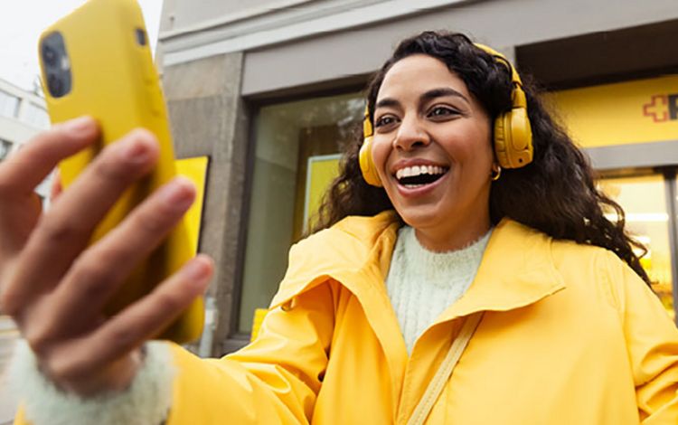 Junge Frau mit gelbem Smartphone und gelber Jacke lacht und macht ein Selfie